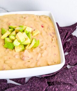 A white square bowl is filled with dairy free queso and topped with diced avocado. Blue corn tortilla chips sit around the bowl.