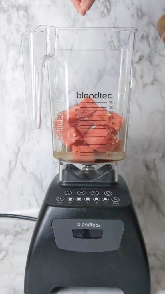A hand adding a pinch of salt to a blender that's partially filled with watermelon.
