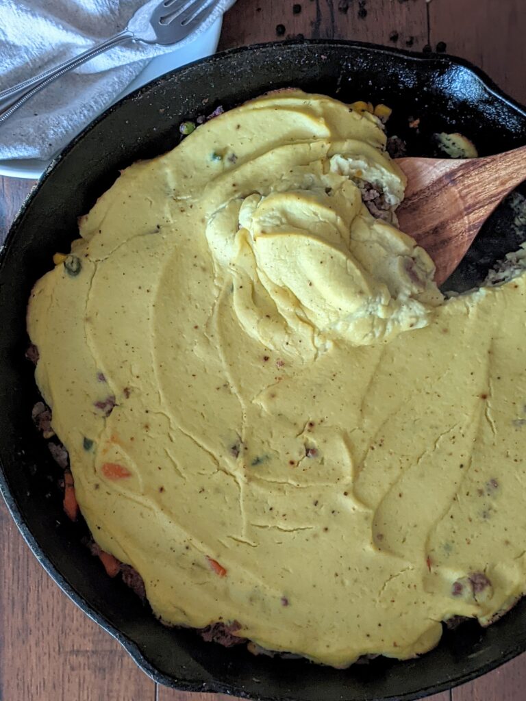Cast iron pan filled with cauliflower shepherd's pie. A wood spoon is starting to dig into the pie. 