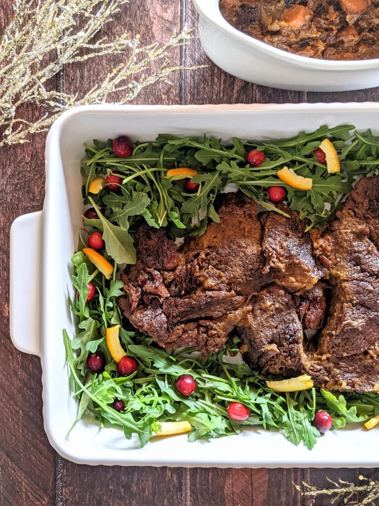Half casserole dish filled with roast, surrounded by arugula, cranberries, and orange peel.