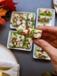 A hand holds a celery bite that's filled with cashew cream, and topped with green onion and bacon bits.
