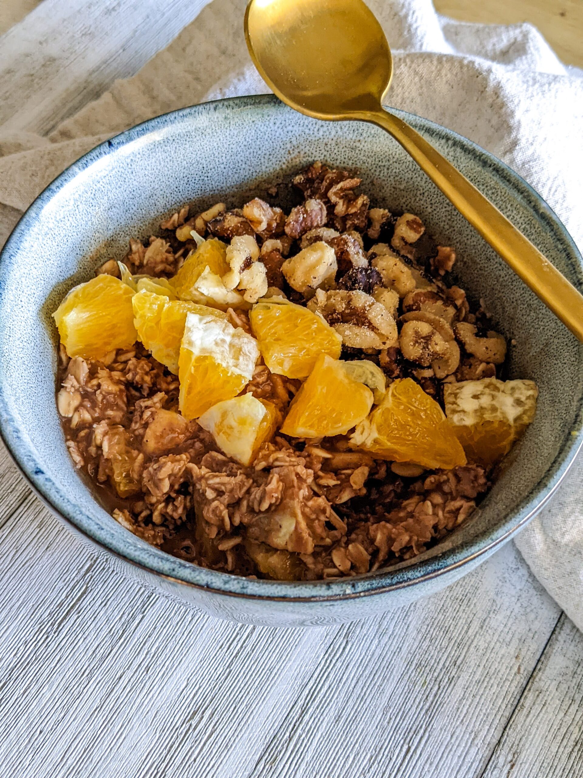 Bowl of chocolate orange oatmeal, topped with chopped walnuts and diced oranges. A gold spoon is laying diagonally across the top of the blue bowl.