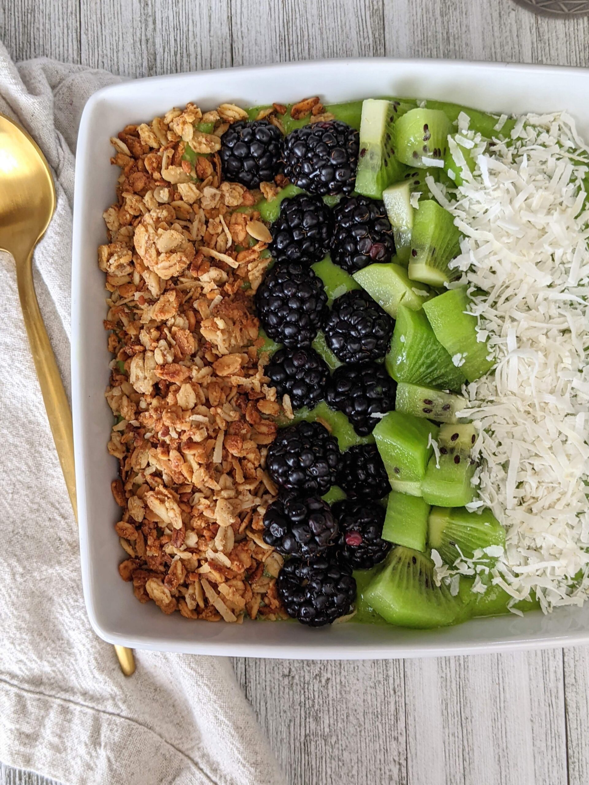 White square bowl filled with green smoothie, topped with granola, blackberries, kiwi, and coconut shreds.