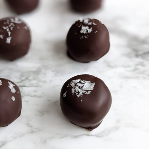 Closeup angle of the nutty bon bons sitting on white marble. Each ball is evenly spaced about an inch apart and each is topped with flaky sea salt.