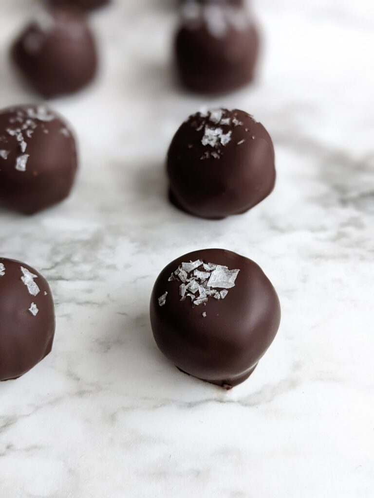 Closeup angle of the nutty bon bons sitting on white marble. Each ball is evenly spaced about an inch apart and each is topped with flaky sea salt.