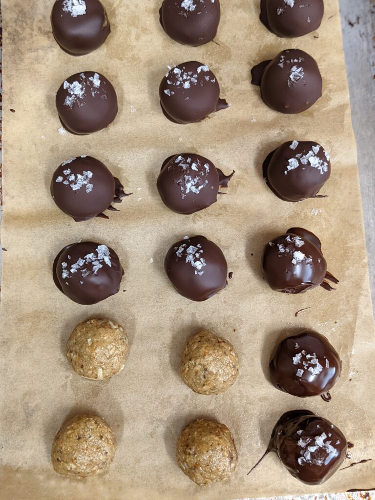 Bon bons on parchment paper. All but 4 are covered in chocolate and topped with sea salt, showing the process.