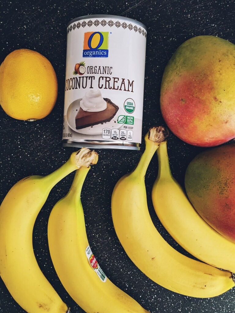 Ingredients to make the mango banana nice cream laid out on a black cutting board. Bananas, lemon, coconut cream, and mango.