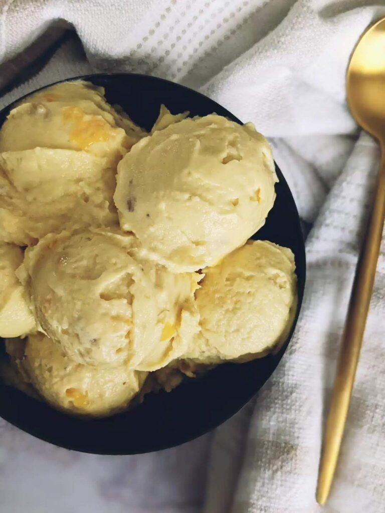Top view of a black bowl filled with scoops of mango banana nice cream. A gold spoon sits on a cloth next to the bowl.