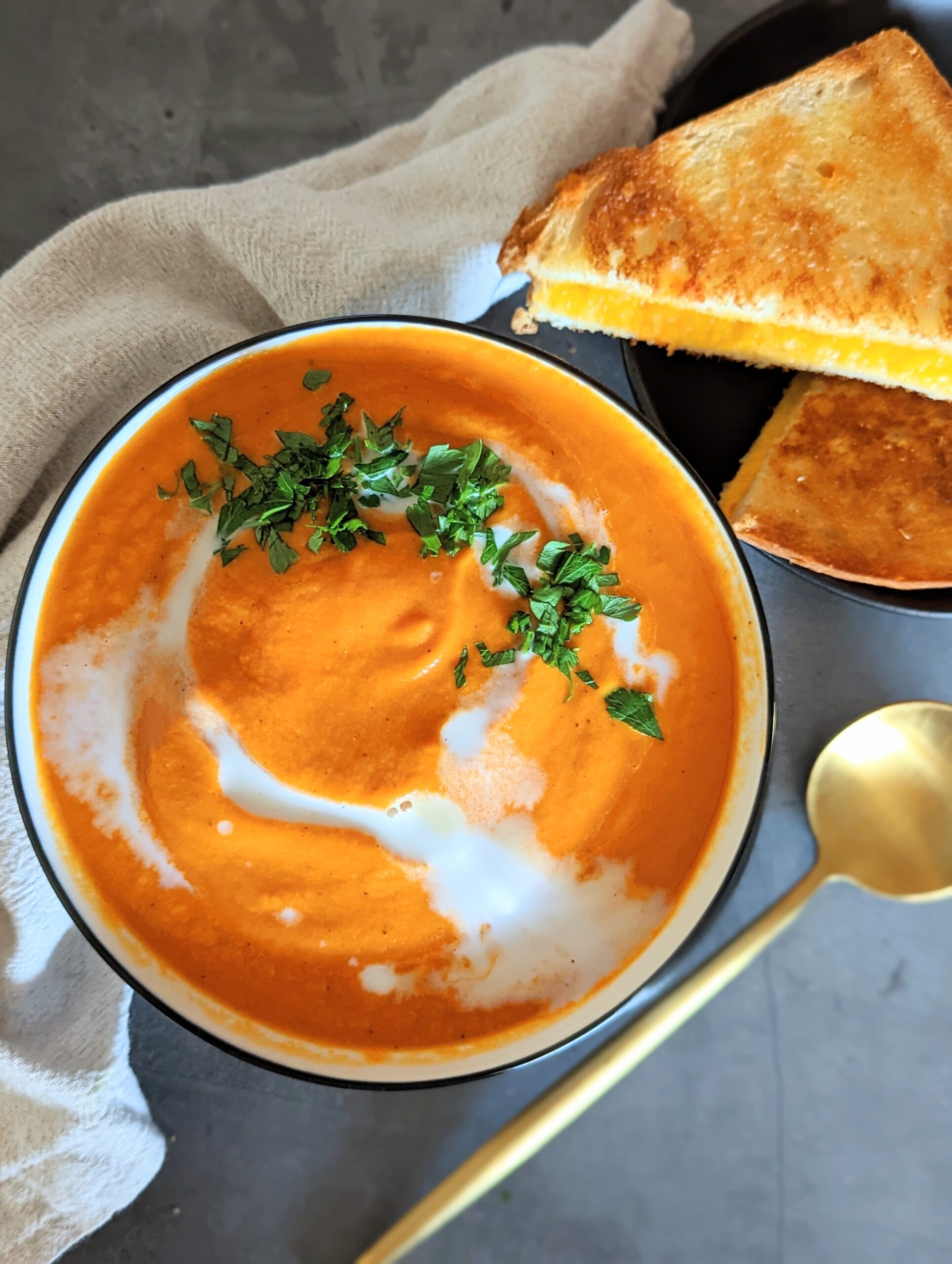 Top view of a full bowl of tomato soup, with swirled coconut cream and chopped parsley on top. A plate of grilled cheese sits next to the bowl.