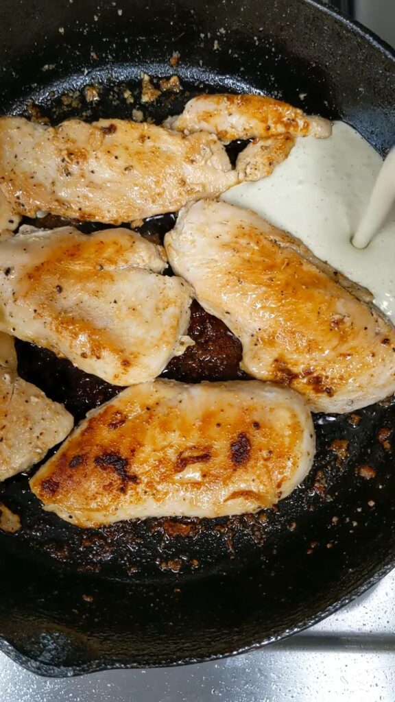 A cast iron skillet has cooked chicken and a creamy sauce is being poured into the pan.