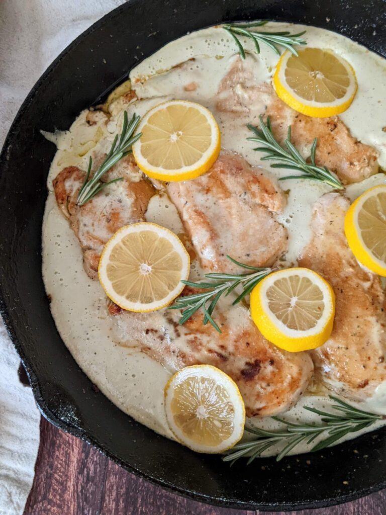 A cast iron skillet holds cooked chicken breasts surrounded by creamy sauce, topped with lemon slices and rosemary sprigs. 