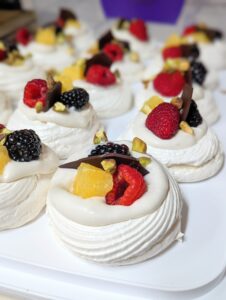 Fully assembled mini pavlovas. Each is topped with cream, one raspberry, one blackberry, one orange piece, chopped pistachios, and a shard of chocolate.