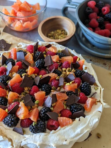 A large fully assembled pavlova with bowls of berries, oranges, and pistachios behind it.