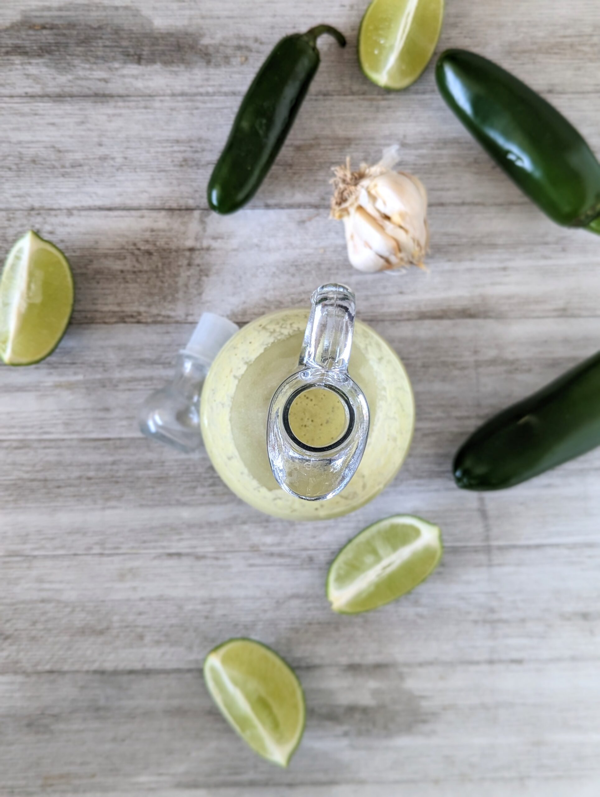 Overhead shot of a glass dressing pourer that's filled with the jalapeno vinaigrette. Lime wedges, a garlic bulb, and jalapenos surround the glass.