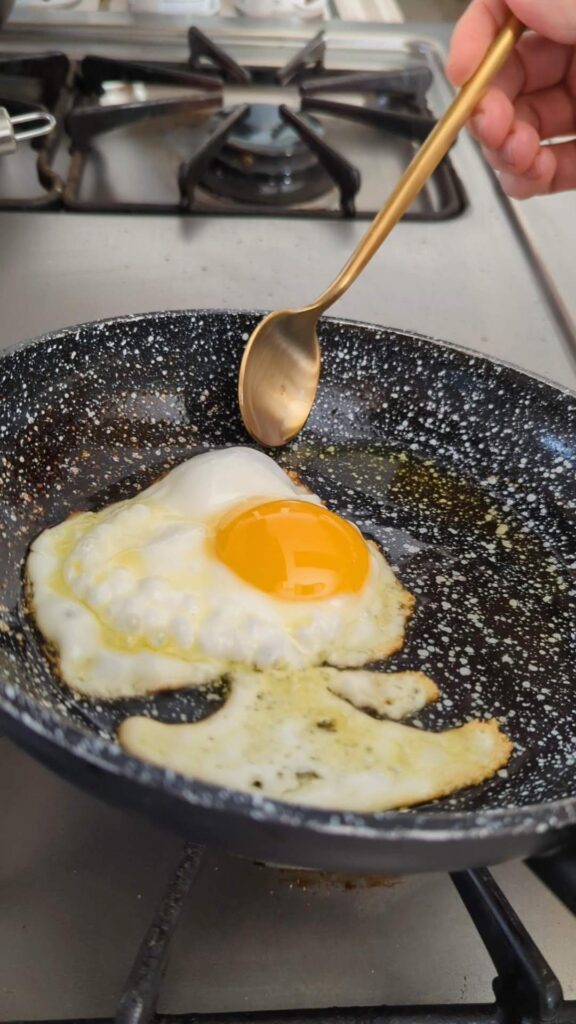 An egg is frying on a skillet and a gold spoon is gathering some of the oil on the pan to pour over the egg.