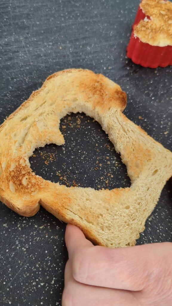 A piece of toast is on a cutting board. A heart shape is cut out of the center.