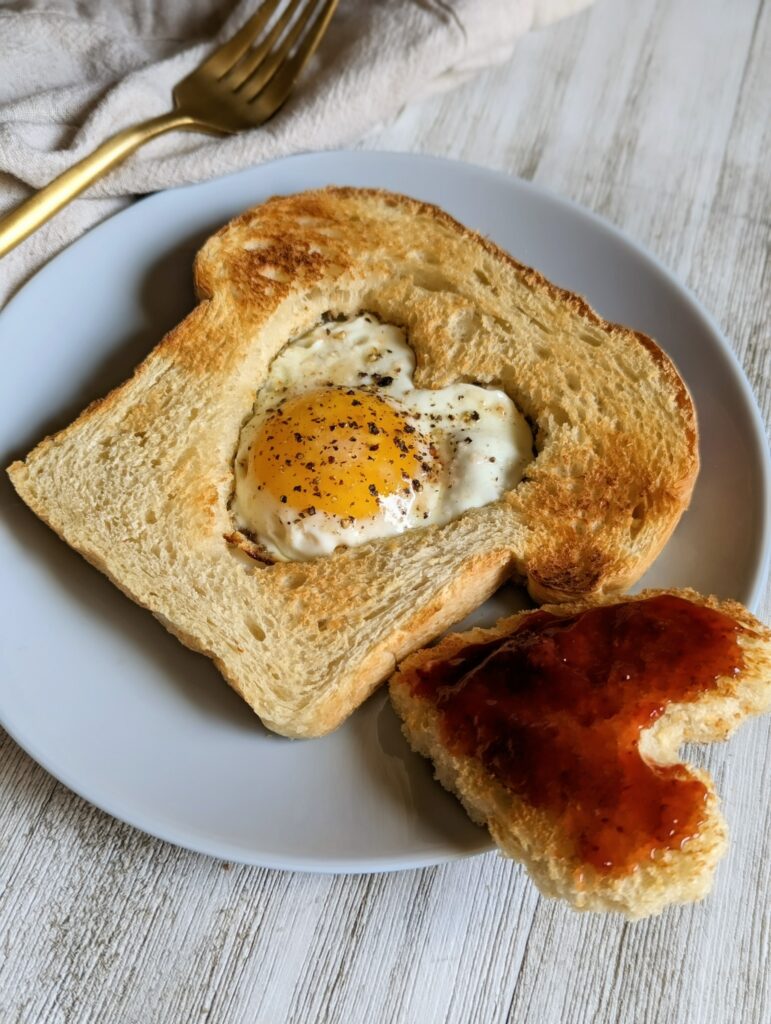 The toast with the heart shaped egg in a hole is on a plate. The center heart piece that was cut from the toast is on the side with strawberry jam on it.