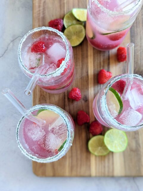 Overhead view of cups of the sparkling raspberry lemon limeade, surrounded by slices of lime and raspberries.
