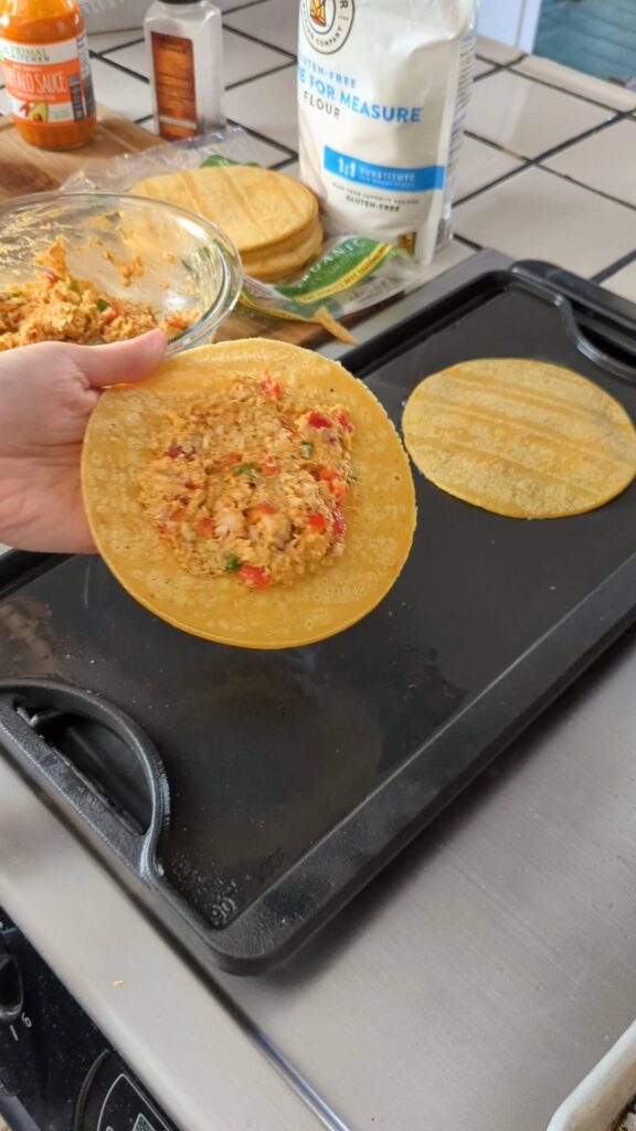Holding an uncooked smash taco over the griddle. The mix in spread on the center of the corn tortilla, and the person is about to flip it face-down onto the griddle.